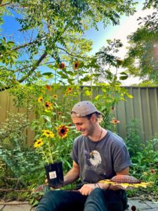 Josh is in his backyard holding and looking at a sunflower. Sunflowers in the backrgound.